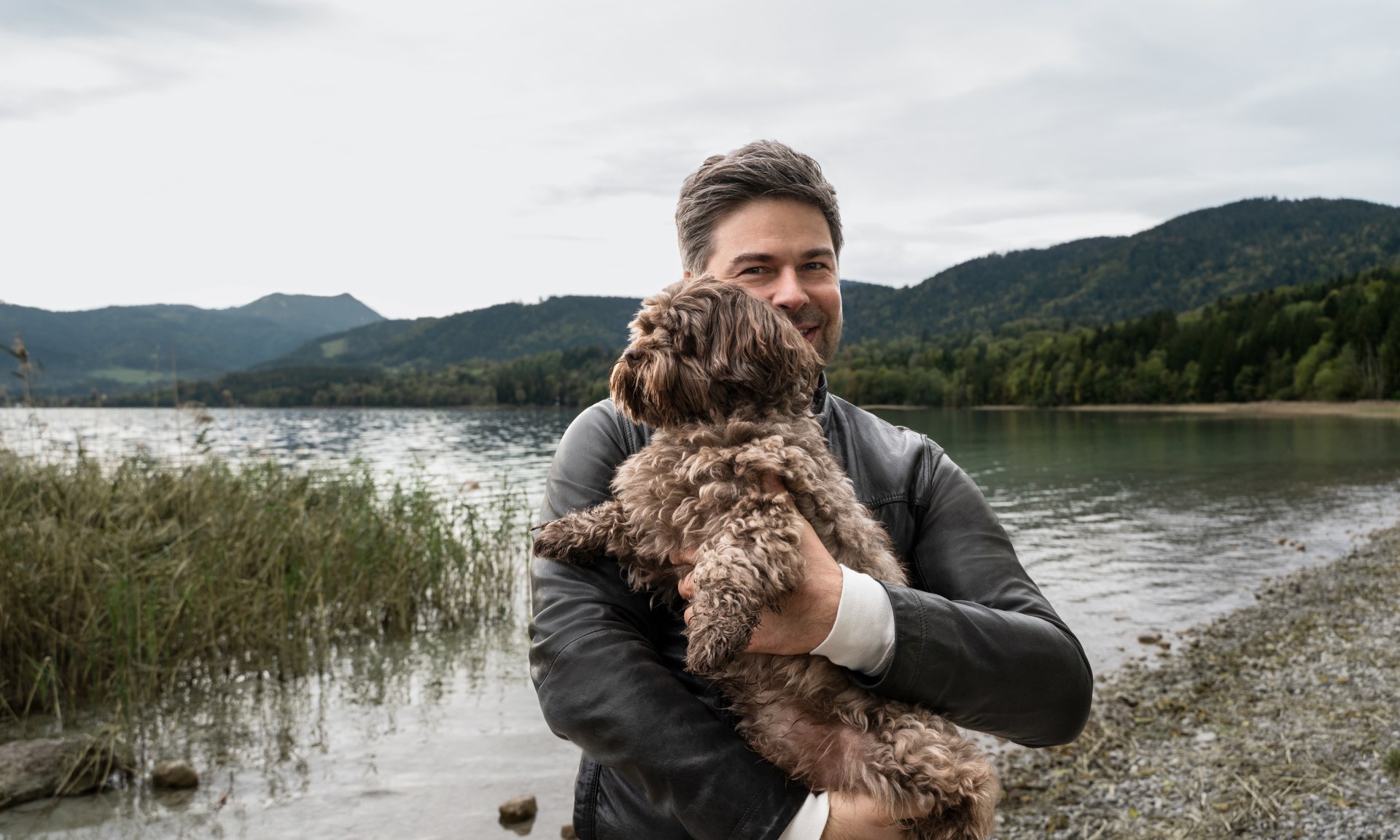 Nils Wollny stoi nad jeziorem z psem na rękach.